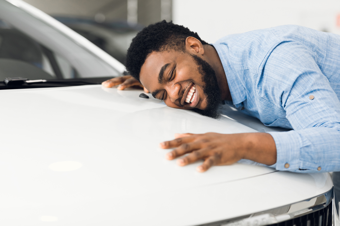 Happy African American Man Hugging New Car In Auto Dealership